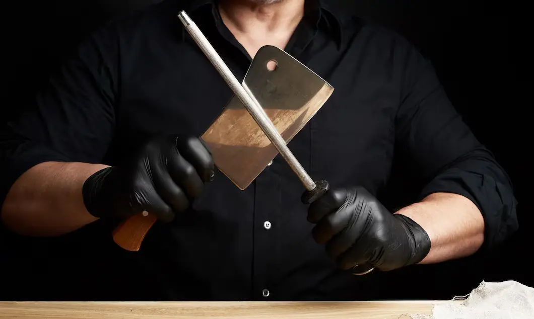 Eine Person in schwarzer Kleidung und schwarzen Handschuhen schärft ein grosses Hackmesser mit einem Wetzstab. Das Messer hat einen Holzgriff, und die Szene findet vor einem dunklen Hintergrund statt, was den Fokus auf das Messer und den Schärfvorgang lenkt.