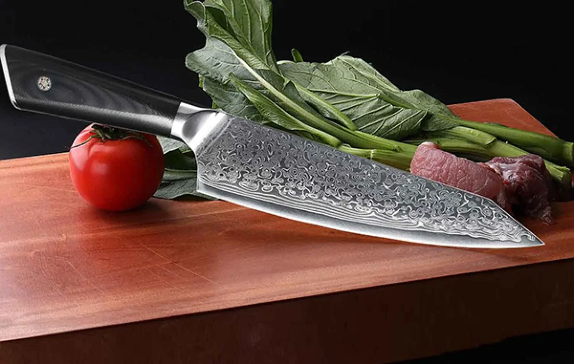 Traditionelles Damastzener Edelstahlmesser gallery 1 Ein Damastmesser liegt auf einem Holzschneidebrett neben einer frischen Tomate, Blattgemüse und einem Stück rohem Fleisch. Die Szene ist bereit für die Zubereitung eines Gerichts mit frischen Zutaten.